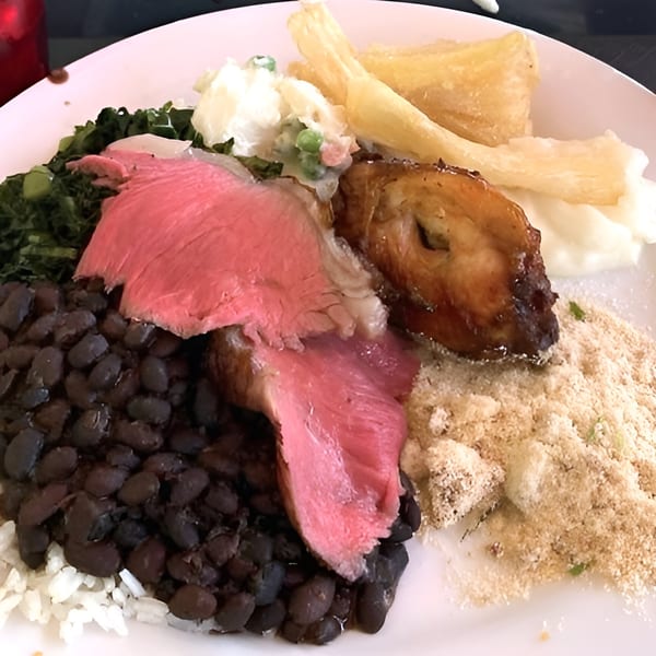 A Brazilian plate with sliced steak, black beans, rice, farofa, collard greens, and fried yucca.