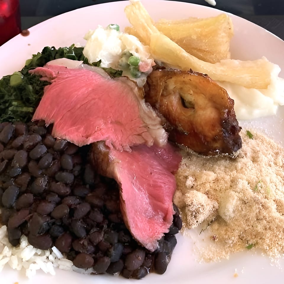 A Brazilian plate with sliced steak, black beans, rice, farofa, collard greens, and fried yucca.