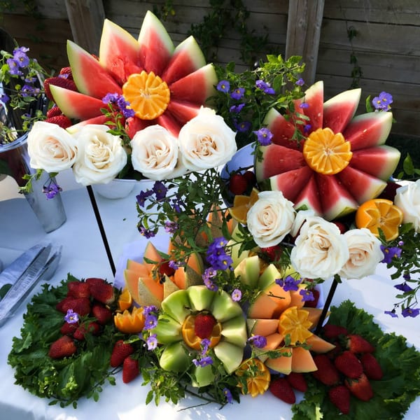 Beautifully arranged fruit platters with watermelon, cantaloupe, strawberries, and oranges, garnished with white roses and purple flowers.