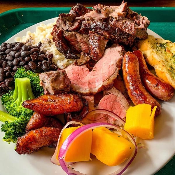 A Brazilian plate with various grilled meats, black beans, rice, broccoli, plantains, and garlic bread.