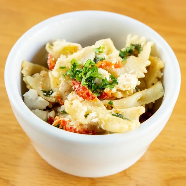 A white bowl filled with farfalle pasta salad, sun-dried tomatoes, feta cheese, and parsley.