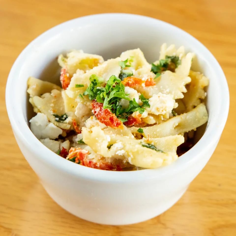 A white bowl filled with farfalle pasta salad, sun-dried tomatoes, feta cheese, and parsley.