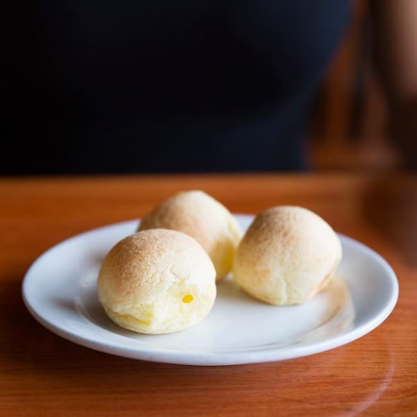 Three golden-brown Brazilian cheese breads (Pão de Queijo) on a white plate on a wooden table.