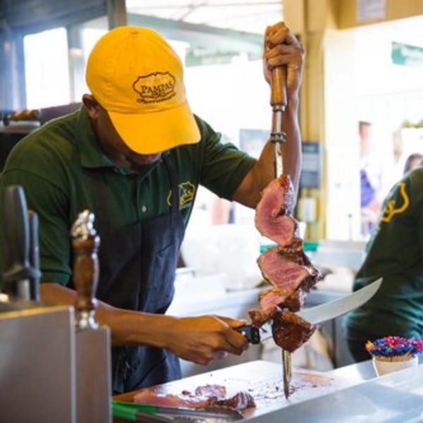 A Pampas Grill chef in a yellow hat carves Brazilian picanha steak from a large skewer.