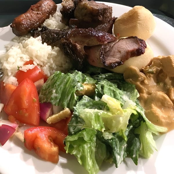 A Brazilian plate with rice, grilled meats, bread, tomato salad, green salad, and a creamy sauce.