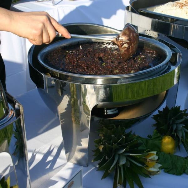 A hand serving black beans from a chafing dish, with pineapple tops and lemon garnishes.