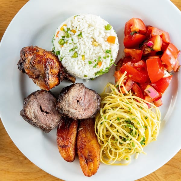 A Brazilian plate with grilled chicken, steak, rice, tomato salad, fried plantains, and pasta.