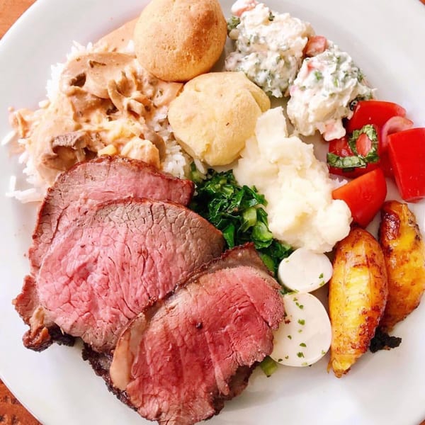 A Brazilian plate with sliced beef, rice, mushroom stroganoff, cheese bread, potato salad, mashed potatoes, and roasted potatoes.